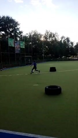 The video shows a group of children playing soccer on an artificial turf field. The children are dressed in athletic wear and are kicking a soccer ball around. There are several tires on the field, which appear to be used as obstacles. In the background, there are trees and a fence. The sky is clear and there are some clouds. The sun is shining brightly.