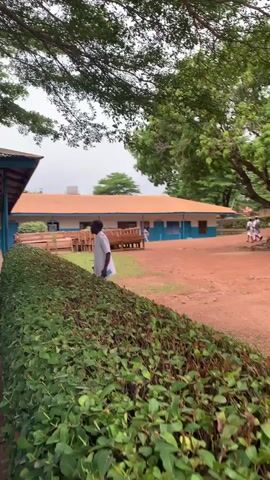 A video shows a schoolyard with a building in the background and trees on either side. In the foreground, a trimmed green hedge lines the bottom of the frame. A person in a white shirt and dark pants is standing near the hedge on the left, facing away from the camera. In the middle ground, a few people in white uniforms are walking across a dirt path towards the right side of the frame. A white bus is parked on the left side of the frame. The building in the background appears to be a single-story structure with a light-colored roof and blue trim. Several trees with green leaves are visible throughout the scene.