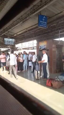 The video shows a busy train platform. People are standing, walking, and sitting, waiting for trains. Some have luggage. A digital display shows train information. There are signs in English and another language. The platform is covered and shaded.