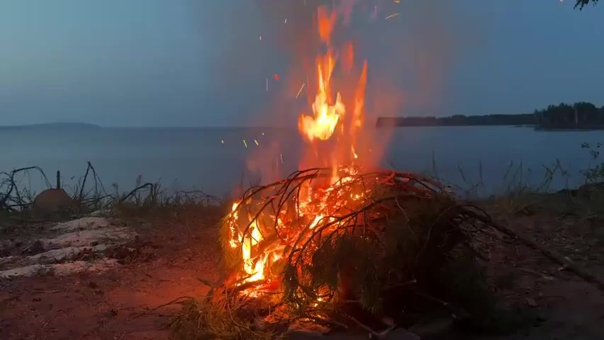 A campfire burns on the shore of a large body of water. The fire is made up of branches and green pine needles, and flames are leaping high into the air. Sparks are flying from the fire, and smoke is rising. The sky is dark, and the water is calm. In the distance, there are trees on the far shore. The ground around the fire is covered in dirt and some scattered rocks. There is a metal object, possibly a pipe, to the left of the fire. The scene suggests a natural, outdoor setting, possibly for camping or relaxation.