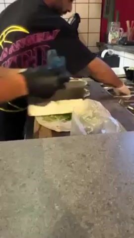 Two men wearing black gloves are preparing burritos on a counter. They are placing ingredients onto tortillas. The first man is wearing a black t-shirt with the word "ANGELS" written in pink and yellow. The second man has a beard and is wearing a black shirt. There is a plastic bag with lettuce on the counter. There is a tiled wall behind the counter. There is a small cactus decoration on the counter in the background.