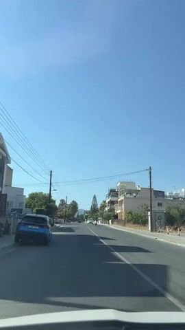 The video shows a view from inside a vehicle, looking down a paved road on a clear, sunny day. The road is lined with trees and buildings on both sides. There are power lines running overhead. A blue car is parked on the left side of the road. A person on a scooter is visible in the distance, riding on the right side of the road. The sky is a clear blue.