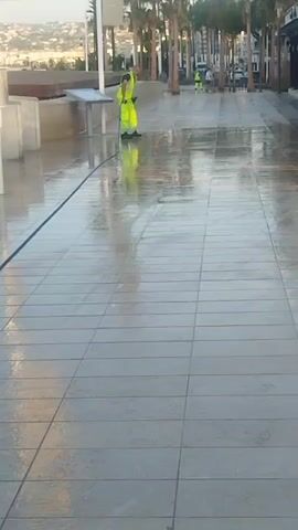 The video shows an outdoor tiled walkway with palm trees in the background. A person wearing a high-visibility yellow vest and green pants is cleaning the walkway with a hose, spraying water. The walkway is wet and reflects the surroundings. Another person in a similar vest is visible further down the walkway. A woman is running towards the camera on the left side of the walkway. The scene appears to be a public area, possibly a promenade or boardwalk.