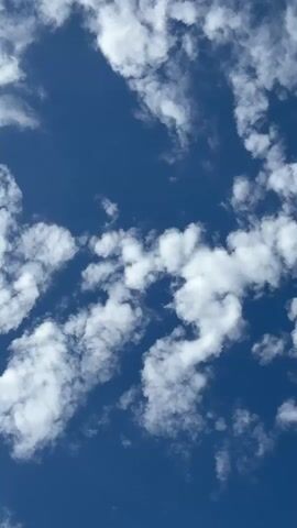 This is a vertical, close-up shot of a bright blue sky with scattered white clouds. The clouds are wispy and appear to be moving slowly across the sky, casting soft shadows. The lighting suggests it is daytime, likely a sunny afternoon. There are no people, animals, or man-made objects visible in the frame. The focus is entirely on the natural beauty of the sky.