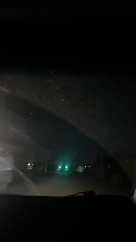 The video captures a dark, nighttime scene from inside a vehicle. The headlights of the car are illuminating a dusty road ahead. In the distance, several traffic lights glow green. The sky is black and appears to have a faint, greenish haze. The overall impression is one of driving through a quiet, possibly rural area at night.