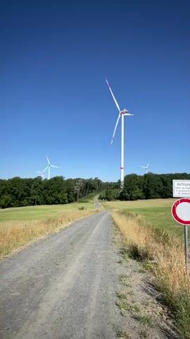 This is a vertical, first-person perspective video of a dirt road leading through a rural landscape with wind turbines. The scene is set in Berfa, Germany, under a clear blue sky, suggesting a sunny day. The road is flanked by fields of dry, yellow grass and lush green trees. In the distance, several tall white wind turbines with red accents are visible, their blades slowly rotating, indicating the presence of wind.

The camera is moving forward, giving a sense of travel along the road. The focus is sharpest on the foreground road and the nearest wind turbine, with the background becoming slightly softer. A sign with German text is partially visible on the right side of the frame, but the text is not legible. The overall mood is serene and natural, highlighting the interaction between man-made technology and the environment. The time of day appears to be mid-day or early afternoon due to the bright sunlight and clear sky. No people are visible in the video.