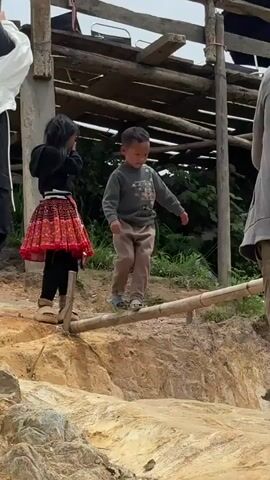 The video shows two children and an adult outdoors. A young boy in a gray sweatshirt and tan pants is walking across a bamboo pole. A younger girl wearing a black hoodie, a red and black patterned skirt, and black leggings stands on the left, looking at the boy. An adult in a gray hooded sweatshirt and tan pants is walking away from the camera on the right. The background features a rustic wooden structure with a corrugated metal roof, lush green foliage, and a dirt embankment. The ground is uneven and appears to be made of soil and rocks.