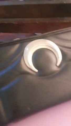 The video shows a close-up of a black, quilted leather bag with a large, silver crescent moon emblem in the center. A metallic chain strap is visible on the bottom left. The bag is resting on a light purple surface and the edge of a dark wooden structure is partially visible at the top. Music plays in the background.