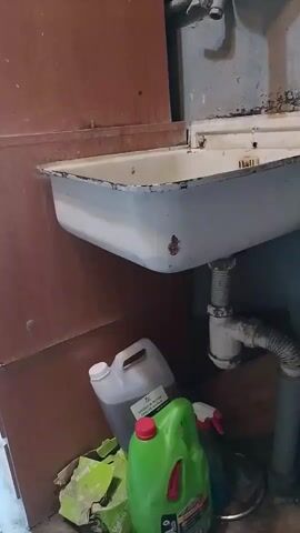 The video shows a utility sink with a metal basin and a faucet. Below the sink, there are several cleaning products, including a large green bottle with a red cap and a clear jug with a white cap. There are also some discarded cardboard boxes on the floor. The walls are a muted brown and grey color. The video is very brief and doesn't show any people or actions.