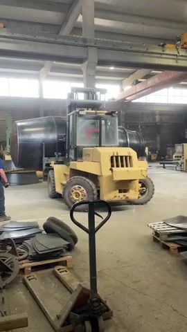 The video shows an industrial workshop or factory interior. A large, yellow Hyster forklift is the central focus, slowly moving a substantial, dark, cylindrical object (likely a tank or pressure vessel). A man wearing a blue t-shirt and jeans walks alongside the forklift, appearing to monitor or guide its movement. Another individual is visible inside the forklift's cab, operating the machinery.

The setting is a spacious industrial building with high ceilings and concrete support pillars. Natural light streams in from large windows positioned high on the walls, indicating it is daytime. Overhead cranes are visible in the background, further emphasizing the industrial environment. The concrete floor is scattered with various metal components, including stacks of sheet metal on wooden pallets and a black pallet jack in the foreground. The brand name "HYSTER" is clearly visible on the side of the forklift. The scene depicts a routine heavy-lifting and transportation operation within the facility.