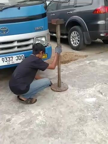 A person is crouching next to a blue truck and operating a jack stand. The person is wearing a black shirt, blue jeans, and gloves. The jack stand is made of metal and has a wide base. The truck is parked on a concrete surface. In the background, there is a dark-colored SUV. The scene appears to be an outdoor setting.