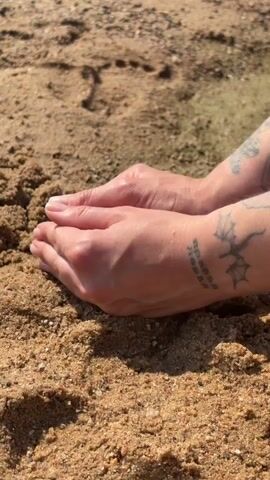 The video shows a close-up of two human hands on a sandy surface. The hands are cupped together at the beginning of the clip. A small butterfly lands in the cupped hands, its wings are dark with some reddish-brown markings. The butterfly is still for a moment before the hands gently open and move away, releasing the butterfly. The scene is set on a sandy beach, likely near a body of water, as evidenced by the rippling water and wet sand visible in the background. The lighting suggests it is daytime, possibly late morning or afternoon, with clear sunny weather. The person's arms have tattoos, including what appears to be a dragon-like creature and some text, though it's too blurry to read. The overall mood is one of gentleness and care towards nature.