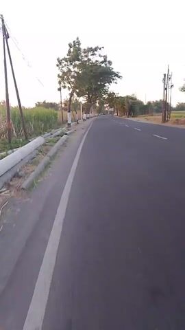 The video shows a road with a person riding a scooter. The road has a white line marking its edge. There are trees and vegetation on the left side of the road. The sky is visible and appears to be a light color, possibly indicating early morning or late afternoon. There is another scooter in the distance.