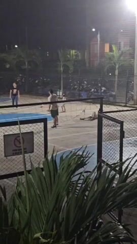The video shows a group of people playing what appears to be volleyball on an outdoor court at night. The court is surrounded by a chain-link fence and vegetation, including palm trees. There are several individuals on the court, including adults and children, engaged in the game. One person is lying on the ground near the net. A sign is visible on the fence, written in Spanish. In the background, there is a parking area with motorcycles and some buildings. The lighting is provided by overhead lamps, creating a somewhat dim environment.