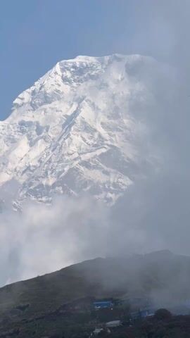 The video captures a majestic, snow-capped mountain dominating the upper two-thirds of the frame under a clear blue sky. The mountain's rugged slopes are heavily covered in white snow and ice, with visible rock faces protruding through the snow. Wispy, white clouds partially obscure the lower sections of the peak and drift across the mid-ground.

In the foreground, a dark green ridge or smaller hill is visible, with some dark-colored structures, possibly buildings or shelters, scattered along its crest. A faint outline of what appears to be a railing or viewing platform is also discernible on the ridge. Throughout the video, a single bird can be seen soaring briefly in the sky near the mountain peak around the 0:03 mark.

The weather appears partly cloudy but bright, with good visibility of the distant peak. The setting suggests a high-altitude, mountainous region, consistent with the provided context of Lwang Ghalel, Nepal. The video is a static shot, likely taken from a viewpoint, capturing the grandeur of the landscape. Human voices speaking in what sounds like Nepali are heard in the background throughout the recording. No visible text is present in the video.