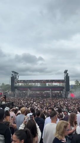 The video captures a wide shot of a large outdoor concert or festival. A massive crowd of people, densely packed, extends from the foreground to the stage in the distance. The crowd is diverse in age and attire, with many individuals holding up their phones to record or take pictures. Dominating the background is a large, elaborate stage setup featuring multiple speaker towers and a wide screen displaying visuals. Smoke or fog drifts across the stage and over the crowd, adding to the atmosphere. The sky above is overcast with dramatic, dark clouds, suggesting the possibility of rain or simply a dramatic, cloudy day. The overall impression is one of a lively, energetic event with a significant turnout.