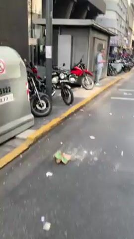 The video shows a street scene in a city, with parked motorcycles lining the curb.  A man stands next to a gray booth, looking at a phone or tablet.  Across the street, a building with a restaurant called "La Panera" is visible.  Traffic passes on the street.