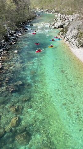 The video shows a group of people kayaking down a clear, turquoise river. There are about 8 kayakers, most of whom are in red or yellow kayaks. The river flows through a rocky, forested area. The water is so clear that the rocks at the bottom are easily visible.  The kayakers are spread out along the river, navigating the mild rapids. The perspective is from above, likely from a bridge or high vantage point. The day appears to be sunny and the overall scene is calm and peaceful.