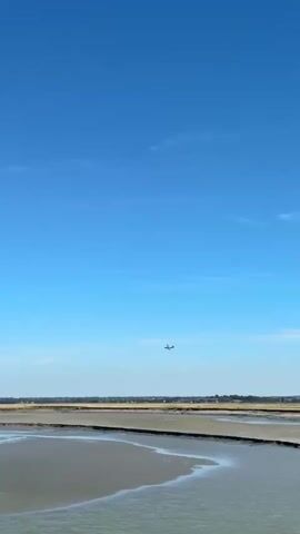 The video shows a vast, clear blue sky with a few wispy clouds. In the distance, a small airplane is visible flying horizontally across the frame. Below the sky, there is a landscape of dry, cracked earth with lines and textures, suggesting it may be a dried-up riverbed or salt flats. The foreground features a body of water, possibly a calm sea or a large pond, with a slightly rippled surface reflecting the light. The horizon line is low, emphasizing the expanse of the sky and the open landscape. The overall impression is one of a bright, sunny day in a spacious, natural environment.