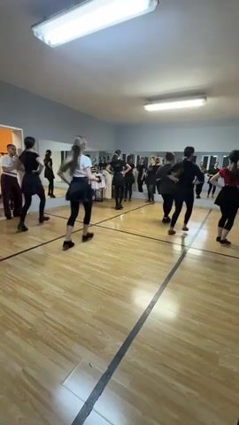 A dance rehearsal is taking place in a well-lit dance studio with a wood floor marked with dark lines, likely delineating practice areas. A group of approximately ten young women, mostly dressed in black and white dance attire, are performing a synchronized dance routine. A man wearing casual clothing is standing near a doorway, observing the dancers. A large mirror covers one wall, reflecting the dancers and the studio. Fluorescent lights are visible on the ceiling. A camera is present, possibly hand-held. The video is likely taken from the corner of the room.