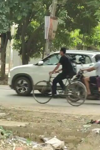 The video shows a street scene with several vehicles and trees. In the foreground, there is a dirt path with some debris. In the middle ground, a person on a bicycle rides past a motorcycle with two people on it. A white SUV is seen in the background on the left. Trees line the street, providing shade. The overall environment appears to be an urban or suburban area with some vegetation.