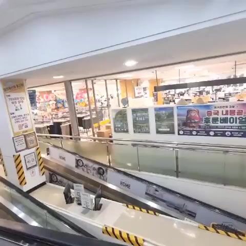 The video shows an interior view of a shopping mall with escalators. In the foreground, a metal railing of the lower escalator is visible. The escalator steps are dark grey and metallic, with yellow and black striped safety markings along the sides. Above this, another escalator is seen with white sides and similar yellow and black safety markings. Several people are present on the escalators. On the upper escalator, a person with dark hair is pushing a yellow shopping cart. Further up, another person is lying down. On the lower escalator, several individuals are standing, some wearing masks. In the background, a bright, well-lit store interior is visible, with shelves stocked with merchandise. Large banners with Korean text and images are displayed along the upper level. The overall environment appears to be a busy retail space.