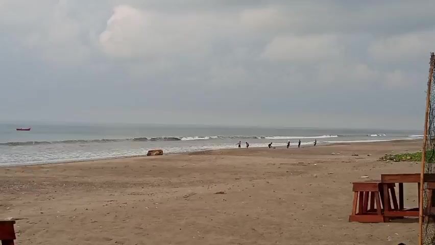 The video shows a beach with people walking on the shore. The sky is cloudy and the ocean has waves. There is a small red boat in the distance. There are also some wooden chairs and a volleyball net visible in the foreground.