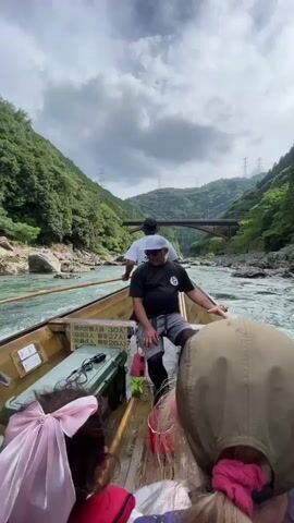 A video shows a long, narrow wooden boat moving down a river surrounded by lush green mountains. The river is rocky and appears to be moving swiftly. A man in a white hat and black shirt is steering the boat from the back, using a long pole to navigate. Another man in a white t-shirt and black hat stands in front of him, also facing the back of the boat. There are several people in the boat, some with their backs to the camera, including a young girl with a pink bow in her hair wearing a life vest. One person in the foreground is holding up a smartphone to record the scene. The sky is cloudy with patches of blue. In the distance, a bridge spans the river, and power lines are visible on the mountainside. The boat has some equipment stored on it, including a green cooler and a red bucket. The water in the river is a murky green color. The overall impression is one of a scenic river journey through a natural landscape.