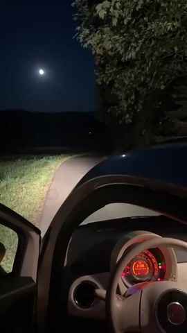 The video captures a nighttime scene from inside a car. The driver's side door is open, and the interior of the car is visible, including the steering wheel and dashboard instruments. The headlights of the car illuminate a paved path that curves into the darkness. The moon is visible in the dark blue sky, and hills are silhouetted against the horizon. Trees with foliage are seen to the right of the frame. The overall impression is one of a quiet, possibly rural, setting at night.