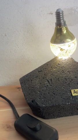 The video shows a table lamp on a wooden table against a white, cracked wall. The lamp's base is a roughly pyramid-shaped black rock with a small gold-colored plate that says "Açores". On top of the rock sits an illuminated, pear-shaped light bulb. A black cable with a black on/off switch is connected to the lamp and lies on the table. The camera angle starts low and slowly moves upwards, keeping the lamp in focus.