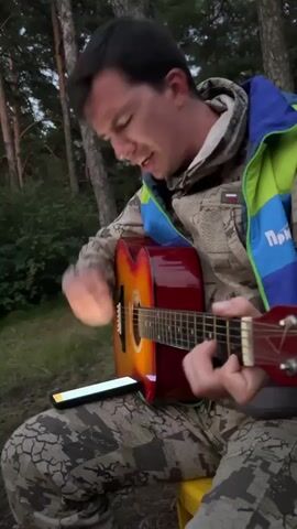 The video shows a group of young people gathered around a campfire in a wooded area. The first segment features a young man playing an acoustic guitar and singing. He is wearing a camouflage jacket over a hoodie. A smartphone is propped up in front of him on the guitar. The second segment shows other young people sitting and watching the guitarist, with a blurry view of the surrounding trees and sky. The third segment shows a wider view of the group sitting in a circle around a campfire, with a person standing and speaking. The final segment focuses on the campfire, with people tending to it and interacting. The overall scene depicts a casual outdoor gathering, possibly a camping trip or a hangout in nature.