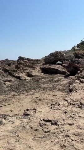 The video shows a rocky coastline under a clear blue sky. In the foreground, there are large, weathered rocks and sandy patches with debris. In the middle ground, the ocean waves crash against the rocks, creating white foam. The background features a green, hilly landscape with scattered white houses and palm trees. The camera pans slightly to the right, revealing more of the coastline and the houses on the hill. No people or specific actions are visible in the video.
