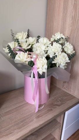 The video presents a medium close-up shot of a bouquet of white roses arranged in a pink cylindrical container. The roses are interspersed with sprigs of green foliage, and the arrangement is partially wrapped in a translucent white paper. Pink and white ribbons adorn the container, adding a touch of embellishment. The bouquet is placed on a wooden shelf with a light, natural finish. In the background, a cream-colored wall and a piece of white furniture are visible, offering a muted backdrop that emphasizes the bouquet's colors and details. The scene is well-lit, showcasing the texture of the roses and the subtle details of the arrangement. Sounds of a child are audible in the background.