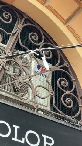 The video shows a close-up shot of an outdoor air conditioning unit mounted behind an ornate metal grate. A small, white, bird-shaped decoration is dangling from a wire, directly in front of the air conditioner. The bird decoration appears to be made of paper or a similar lightweight material and is swaying slightly. The air conditioner itself has a visible fan and a logo that reads "Gree". Below the grate, a black sign with white lettering is partially visible, with the letters "OLOR" clearly legible. The overall scene suggests an urban or commercial setting, possibly an exterior of a shop or building. The lighting is natural, indicating it is daytime.