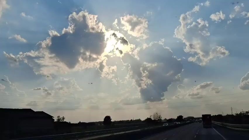 The video captures a view of the sky from a moving vehicle, likely on a highway. The sun is partially obscured by large, fluffy clouds, casting rays of light that create a dramatic effect. The sky is blue with some wispy clouds in the distance. Below the sky, a highway stretches out, with several cars and trucks visible. There are also some trees and buildings on the side of the road. The overall impression is one of a scenic drive on a sunny day.