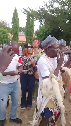 The video shows a small group of people standing on a grassy lawn, interacting with a man in traditional dress. The man is wearing a white shirt, a striped headband, a woven sash, and a decorative fringe of what appears to be animal hair around his waist. To his left, a man in a white t-shirt with a red headband is looking at a paper in his hand. Next to him is a woman wearing a black baseball cap, sunglasses, a floral shirt, and jeans. Beside her is another woman wearing a patterned yellow and green dress and a red headscarf. In the background, more people can be seen milling about, and there are some trees and a building in the distance. It seems like a cultural event or celebration.