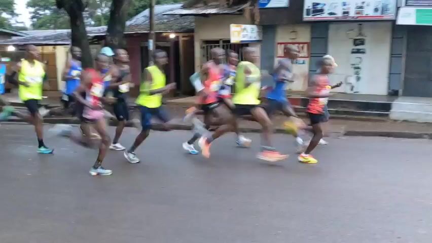 The video shows a group of runners participating in a race or marathon on a paved road in a residential area. The road is lined with simple buildings and trees. Some people on motorcycles and pedestrians are present on the side of the road. The runners are wearing athletic clothing. They are moving quickly along the road. The overall setting suggests a community event taking place in a relatively urban or suburban environment.