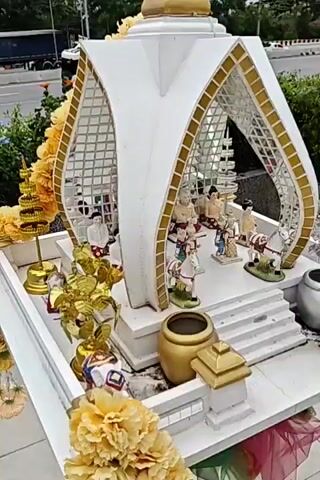 This video pans across an outdoor shrine decorated with white and gold accents. The shrine features numerous small figurines, including several seated figures with elderly features and others dressed in traditional clothing. There are also figurines of horses and what appear to be children. The shrine is adorned with artificial yellow flowers and gold decorative leaves. A large gold pot is visible in the foreground. The background suggests an outdoor setting, possibly near a road, with greenery and what appears to be a truck.