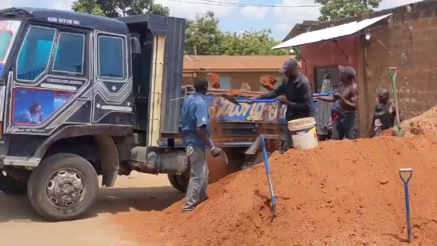 The video shows a group of five men loading red soil into the back of a black truck. One man is shirtless. The truck has the phrases "GOD WITH US" and "BLOOD OF JESUS" written on it. There is an image of Jesus on the truck.  One man is using a shovel to fill a bucket with soil, and another man is helping to lift it into the truck. Two men are digging directly into the soil and tossing it into the truck. One man is sitting. The truck is parked on a street next to a large pile of red soil.  A small building is in the background.