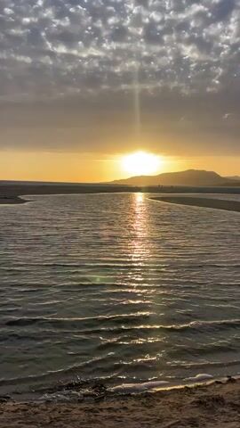 The video shows a sunset over a body of water with a sandy shore in the foreground. The sun is low on the horizon, casting a golden glow across the sky and reflecting on the water. Clouds are visible in the sky, some of which are illuminated by the sun. There are distant hills or mountains in the background. The water appears calm with gentle ripples. A narrow strip of sand separates the foreground from the main body of water. There are a few indistinct figures on the distant landmass. The overall scene is serene and tranquil.