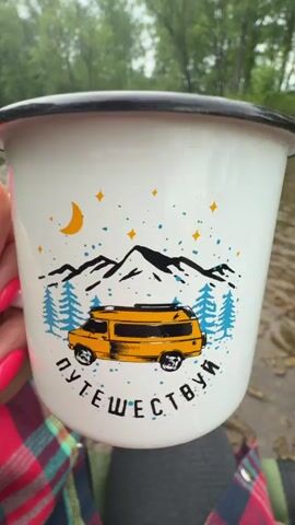 A person sits by a shallow river, holding a white mug with a black rim. The mug is decorated with an image of a yellow van and the words "ПУТЕШЕСТВУИ" (travel) in Russian. Inside the mug is a dark liquid, possibly tea or coffee. The person's hand, sporting bright pink nail polish, holds the mug. They wear gray leggings, black and white striped socks with "NIKE" printed on them, and leopard-print New Balance sneakers. They are also wearing a red plaid shirt. The river is flowing gently, with rocks and mud visible along the banks. Lush green trees line the riverbank in the background. The camera angle is from a first-person perspective, looking down at the person's lap and out towards the river.