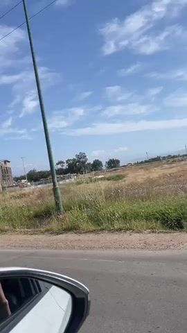 The video shows a car driving past a construction site. The construction site is a large building with a tall tower, likely a mosque, and it is surrounded by a fence. The building is made of brick and concrete, and there is a crane visible on the construction site. The sky is blue with some clouds. The car is moving at a moderate speed. There is a hand visible in the car's side mirror holding a phone.
