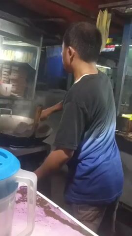 A man is cooking at an outdoor food stall. He is wearing a t-shirt and jeans and is stirring food in a wok with a spatula. Steam is rising from the wok. The stall appears to be open late at night, with a few lights illuminating the area.