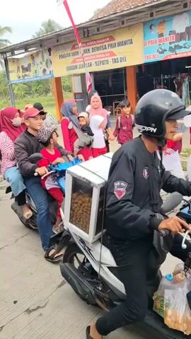 A group of people are participating in a procession. A man wearing a black jacket and a red shirt is riding a motorcycle with a child in front of him. Behind them, a woman in a pink and white striped shirt is sitting on the back of the motorcycle. To the left, a woman in a white and green patterned outfit is riding a white motorcycle with another woman in a matching outfit on the back. A police officer on a black motorcycle is in the lead, and other people in red outfits are walking behind. The procession is taking place on a paved road in front of a building with a banner that has text in Indonesian. The weather appears to be overcast.