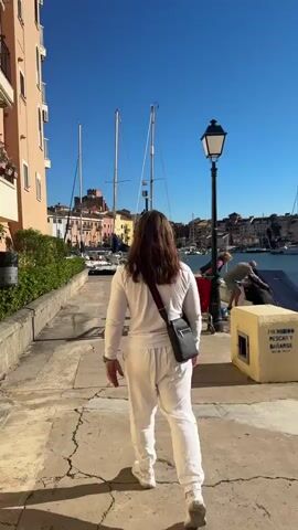 The video shows a waterfront scene on a bright, sunny day. The camera follows a person with long brown hair wearing a white tracksuit and a black crossbody bag as they walk along a paved walkway. To the left is a multi-story building with balconies and green shrubbery. As the camera pans to the right, it reveals a marina filled with various sailboats and yachts. People are seen working on boats, including one couple using a red shopping cart. A young girl in a pink hat and a boy walk along the walkway near a concrete structure with a sign that says "PROHIBIDO PESCAR Y BAÑARSE" (fishing and bathing prohibited). The sun glints off the water, highlighting the boats and the buildings lining the waterfront.