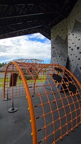This media file showcases an outdoor adventure park in Kamenka, Russia. The scene features a vibrant orange net climbing structure in the foreground, likely part of a children's play area or an obstacle course. Behind it, a tall, grey and white climbing wall dominates the frame, adorned with numerous colorful climbing holds of various shapes and sizes. The wall is designed with a mountain motif, with geometric patterns suggesting peaks and valleys. Climbing ropes are visible, some attached to the wall and others hanging loosely.

The setting appears to be under a covered structure, indicated by the dark roof with visible beams and supports overhead. Beyond the covered area, a bright, partly cloudy sky suggests it's daytime, possibly late morning or early afternoon. The weather seems pleasant, with clear blue sky peeking through fluffy white clouds. Green grass and trees are visible in the background, indicating a park-like or recreational environment.

While no people are clearly identifiable as the main subjects, a person can be faintly seen on the climbing wall, suggesting the facility is in use. The overall impression is one of an active and engaging outdoor space designed for recreation and physical activity, possibly for families or groups. The media file captures a static view of the park, highlighting the equipment and the outdoor environment. There is no visible text in the frame.