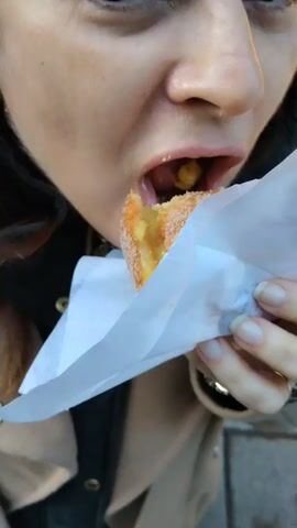 The video captures a close-up view of a fair-skinned woman eating a pastry. She is holding the pastry, partially wrapped in white paper, with her right hand. The camera angle is from slightly below, focusing primarily on her mouth and the lower half of her face. She takes a bite of the pastry, and afterwards, crumbs are visible on her lips. She wears a dark jacket with gold-colored buttons and a neutral-toned outer layer, and minimal jewelry is visible, including earrings and a ring. The environment is not clearly discernable, but appears to be outdoors.