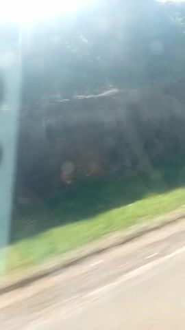 The video shows a view through a car window, possibly a side window, with sunlight glaring and creating lens flares. The background outside the window shows a blurry scene of green vegetation and possibly trees. In the foreground, partly obscuring the view, is the side of a white or light-colored smartphone, specifically the camera module with three lenses visible. The camera appears to be the focal point for a brief moment. The video appears to have been taken while the car is in motion. Raindrops or water spots are visible on the window.