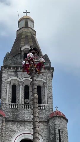 The video shows four people in traditional clothing on top of a tall wooden pole. They are positioned on a small platform at the top of the pole, which has a feathered headpiece attached to it. The pole is situated in front of a large stone church with a tall steeple. The steeple is adorned with a cross at its peak. The sky is blue with some clouds. The people appear to be preparing for a ritual or performance.