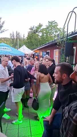 This video shows a crowded outdoor gathering with people dancing and socializing. The environment appears to be a patio or courtyard with some tents or awnings providing shade. There are red buildings in the background and trees and foliage surrounding the area. The lighting suggests it is dusk or evening, with artificial lights illuminating the scene, particularly green lights on the ground. People are dressed in casual summer attire, including dresses, shorts, and t-shirts. Some individuals are interacting, talking, and dancing. A woman in a light-colored, floor-length dress is prominent in the center of the frame, and a man with a beard is seen to her right. Another person, wearing a straw hat with a red ribbon, is partially visible in the foreground. The atmosphere seems festive and lively.